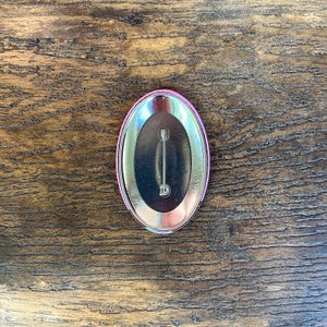 Puede incluir: Un bot&oacute;n ovalado con una parte trasera de metal plateado y un borde rosa y plateado. El bot&oacute;n tiene un cierre tipo imperdible en la parte trasera. El bot&oacute;n est&aacute; sobre un fondo de madera.