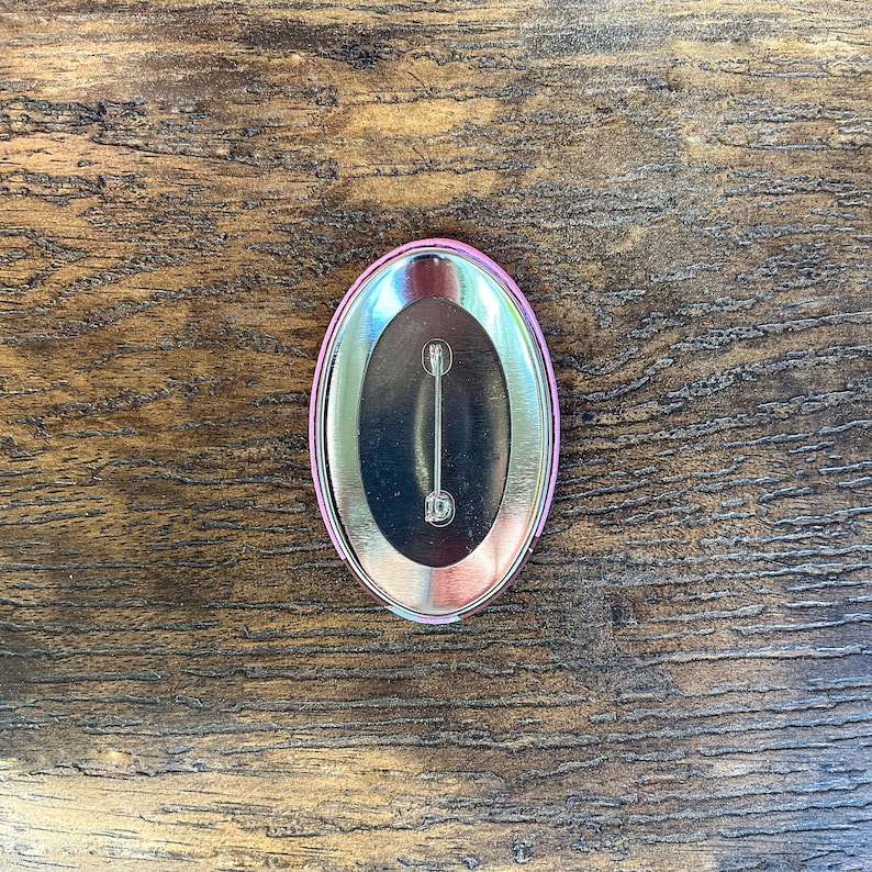 Puede incluir: Un bot&oacute;n ovalado con una parte trasera de metal plateado y un borde rosa y plateado. El bot&oacute;n tiene un cierre tipo imperdible en la parte trasera. El bot&oacute;n est&aacute; sobre un fondo de madera.