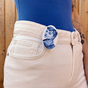 May include: A white oval pin with a blue and white image of Audrey Hepburn. The pin is attached to a person's white jeans.