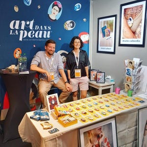 Puede incluir: Un stand de feria con el texto "art dans la peau" en una pared azul decorada con retratos. Una mesa exhibe grabados art&iacute;sticos y un juego con caras ilustradas. Dos personas est&aacute;n detr&aacute;s de la mesa.