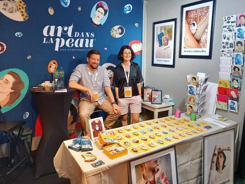 Puede incluir: Un stand de feria con el texto "art dans la peau" en una pared azul decorada con retratos. Una mesa exhibe grabados art&iacute;sticos y un juego con caras ilustradas. Dos personas est&aacute;n detr&aacute;s de la mesa.