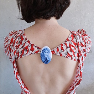 May include: A red dress with a white pattern of hands. The dress has a low back and a white oval-shaped brooch with a blue and white image of a woman's face.