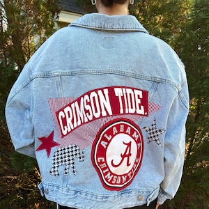 May include: Light blue denim jacket with Crimson Tide patches. Features a red star, a houndstooth elephant, a red and white gingham patch, and a circular patch with the letter "A" and the words "Alabama Crimson Tide".