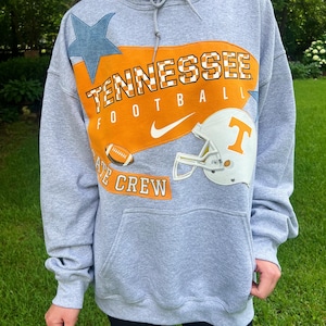 May include: A gray hoodie with a large orange and white graphic that says "Tennessee Football" with a football and helmet. A blue star is on the left shoulder. The hoodie has a drawstring hood and a front pocket.