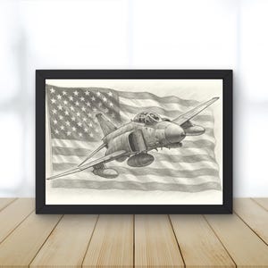 May include: A framed pencil drawing depicting a jet fighter plane in flight, set against the backdrop of the American flag. The artwork is in grayscale, with detailed shading and a black frame. The image is displayed on a wooden surface.