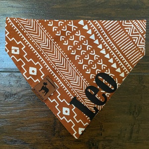 May include: A brown and white patterned dog bandana with the name "Leo" in black text. The bandana has a brown leather tag with a dog silhouette and the text "Muttley Crew Designs".