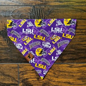 May include: Purple and gold dog bandana with a repeated pattern of LSU Tigers logos and helmets. The bandana has a brown leather tag with a paw print.