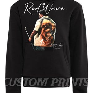 May include: Black hoodie featuring a graphic of a person with a microphone, and the text "Rod Wave" in white script. The hoodie also has the text "M. Lost LP" and "Rod Wave" in white. The words "Custom Prints" are printed at the bottom.