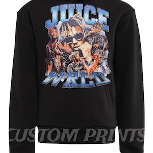 May include: Black hooded sweatshirt featuring a graphic of a musician with the words "JUICE WRLD" in a distressed font. The sweatshirt also has the text "CUSTOM PRINTS" along the bottom.