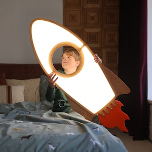 May include: A child holds a wooden rocket ship night light with a bright white light shining through the cutout window. The rocket ship has a red and orange flame design.