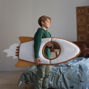 May include: A young boy holds a wooden rocket ship toy with a white, fluffy interior. The rocket ship has a white flame design on the back. The boy is wearing a green hoodie and khaki pants. He is standing in front of a bed with a blue and white patterned duvet cover.
