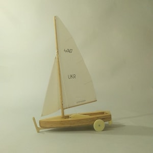 Könnte beinhalten: Ein hölzernes Modell eines Segelboots mit einem weißen Segel. Das Segel trägt die Nummer "470" und das Wort "UKR" aufgedruckt. Das Modell steht auf einer weißen Oberfläche.