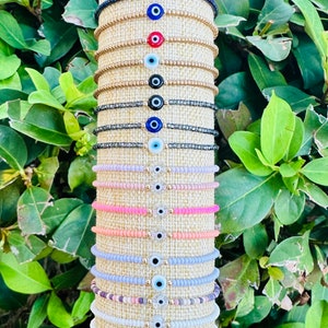 May include: A display of colorful beaded bracelets with evil eye charms. The bracelets are arranged vertically on a beige background. The evil eye charms are in various colors, including blue, red, and black.