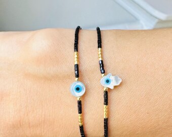 May include: Two beaded bracelets with black and gold beads. One bracelet has a blue evil eye charm and the other has a white hamsa hand charm.