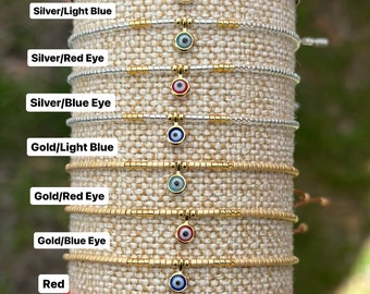 May include: A set of ten beaded bracelets with a gold evil eye charm. The bracelets are in various colors, including black, silver, gold, and red. The evil eye charms are blue, red, and light blue.