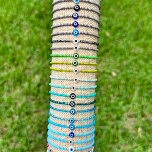 May include: A stack of colorful beaded bracelets with evil eye charms. The bracelets are arranged in a vertical stack on a wooden display stand. The bracelets are made with small beads in various colors, including white, gray, green, blue, turquoise, and pink. Each bracelet features a blue evil eye charm.