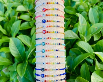 May include: A collection of twenty-one adjustable cord bracelets stacked vertically on a display stand. Each bracelet features a central circular evil eye bead followed by a sequence of small, multi-colored glass beads in a rainbow pattern.