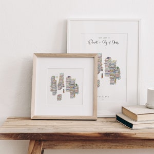 Op de afbeelding: Twee ingelijste illustraties van een stad gemaakt van gestapelde boeken. De tekst op het grotere frame luidt "Get lost in Powell's City of Books, Portland, OR".