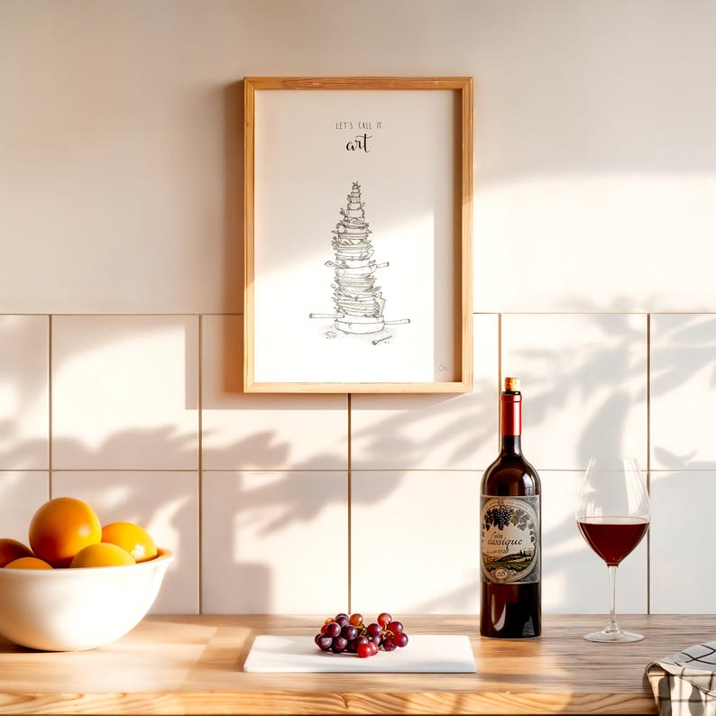 May include: A framed print with a black and white illustration of a stack of dishes and the text "Let's call it art". A bottle of red wine and a glass of red wine are on a wooden table with a white cutting board and a bowl of oranges.