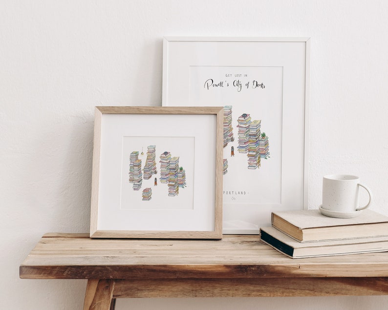 Op de afbeelding: Twee ingelijste illustraties van een stad gemaakt van gestapelde boeken. De tekst op het grotere frame luidt "Get lost in Powell's City of Books, Portland, OR".