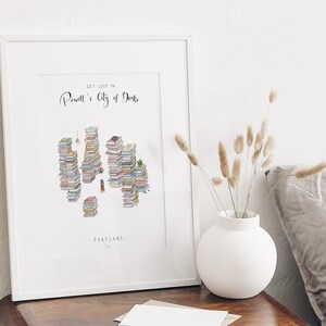 Op de afbeelding: Een wit ingelijst print met de tekst "Get lost in Powell's City of Books" en een illustratie van een stad gemaakt van gestapelde boeken. De tekst "Portland, OR" staat onderaan de print.