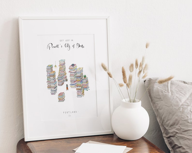 Op de afbeelding: Een wit ingelijst print met de tekst "Get lost in Powell's City of Books" en een illustratie van een stad gemaakt van gestapelde boeken. De tekst "Portland, OR" staat onderaan de print.