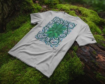 Può includere: Una maglietta grigio chiaro con un vivace design botanico verde e blu. La grafica include un elemento centrale a forma di foglia circondato da viticci e forme di foglie più piccole. La maglietta è esposta su una superficie muschiosa.