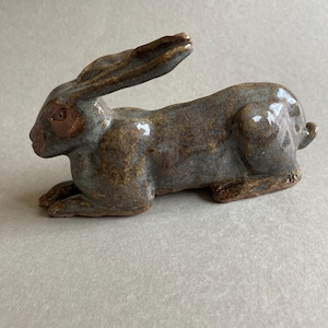 Puede incluir: Figura de conejo de cerámica en posición de descanso. La escultura es principalmente de color gris-marrón con acentos marrón oscuro en las orejas, los ojos y las patas. El conejo mide aproximadamente 20 cm de largo.