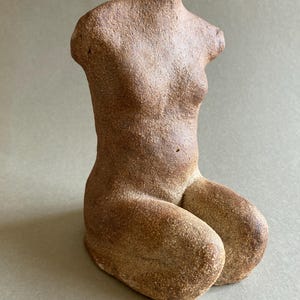 May include: A brown, textured ceramic sculpture of a torso in a kneeling position. The figure is headless and armless, featuring a rough, granular surface finish with earthy tones. The sculpture depicts a seated human form with defined curves.