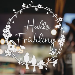Könnte beinhalten: Weißer Vinyl-Aufkleber mit einem floralen Kranz-Design. Der Kranz hat ein Vogelhaus, Vögel und den Text "Hallo Frühling" in der Mitte.
