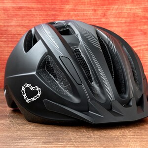 Puede incluir: Casco de bicicleta negro con una pegatina en forma de coraz&oacute;n de eslabones de cadena. El casco tiene orificios de ventilaci&oacute;n y rayas plateadas. Dise&ntilde;ado para la seguridad y protecci&oacute;n durante el ciclismo.