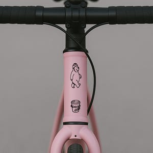 Puede incluir: Un cuadro de bicicleta rosa con manillar negro. El cuadro presenta un dibujo lineal negro de un oso con casco y una taza de caf&eacute;. La bicicleta est&aacute; sobre un fondo gris.