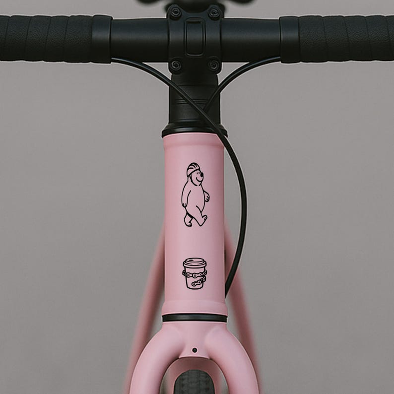Puede incluir: Un cuadro de bicicleta rosa con manillar negro. El cuadro presenta un dibujo lineal negro de un oso con casco y una taza de caf&eacute;. La bicicleta est&aacute; sobre un fondo gris.