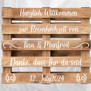 Könnte beinhalten: Ein Holzpalettenschild mit weißem Text, das eine Hochzeitstagfeier ankündigt. Das Schild lautet "Herzlich Willkommen zur Rosenhochzeit von Tina & Manfred Danke, dass Ihr da seid 12. July 2024".