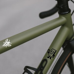 Puede incluir: Primer plano de un cuadro de bicicleta verde oliva con manillar y neum&aacute;ticos negros. Calcoman&iacute;as blancas de una abeja y un paquete de bocadillos sonriente son visibles en el cuadro. La bicicleta tiene un portabid&oacute;n.