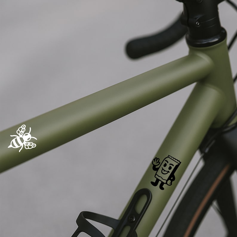 Puede incluir: Primer plano de un cuadro de bicicleta verde oliva con manillar y neum&aacute;ticos negros. Calcoman&iacute;as blancas de una abeja y un paquete de bocadillos sonriente son visibles en el cuadro. La bicicleta tiene un portabid&oacute;n.