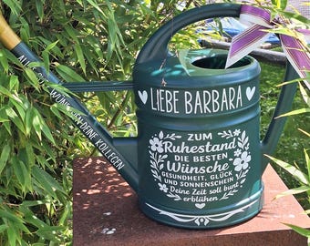 May include: A dark green watering can with a gold spout and a black handle. The can has white lettering that says "LIEBE BARBARA" and "ZUM Ruhestand". A purple and white ribbon is tied to the handle. The can is surrounded by green foliage.