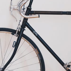 Puede incluir: Una bicicleta negra con manillar plateado y tubo superior envuelto en cuero marr&oacute;n. La bicicleta tiene un logotipo blanco en el cuadro y neum&aacute;ticos negros con llantas doradas. La bicicleta est&aacute; apoyada contra una pared blanca.