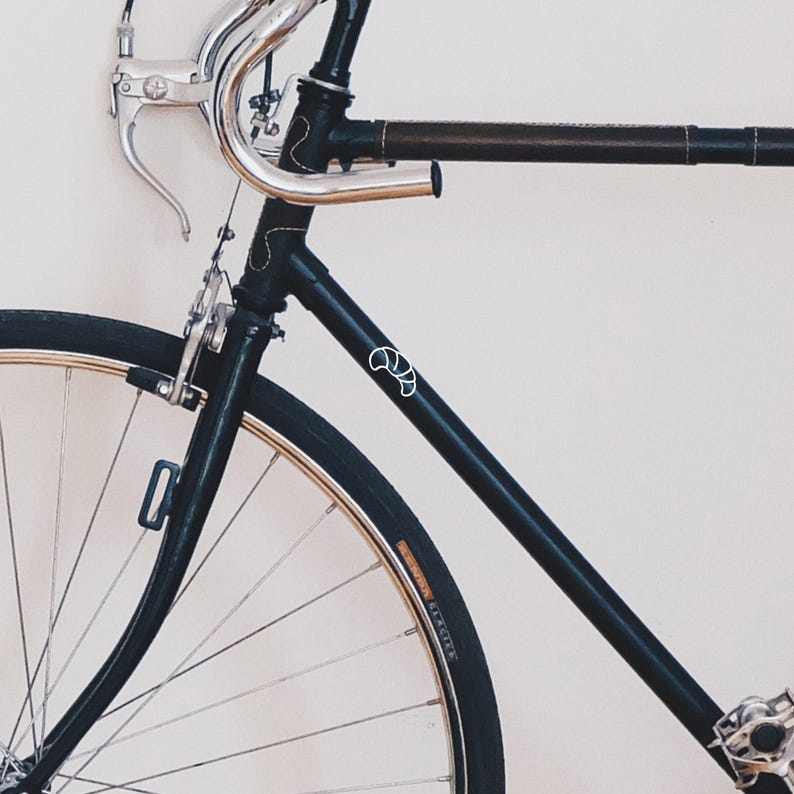 Puede incluir: Una bicicleta negra con manillar plateado y tubo superior envuelto en cuero marr&oacute;n. La bicicleta tiene un logotipo blanco en el cuadro y neum&aacute;ticos negros con llantas doradas. La bicicleta est&aacute; apoyada contra una pared blanca.