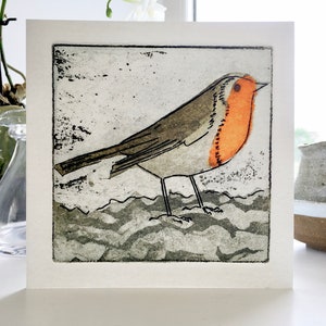 May include: A square greetings card with a print of a robin. The bird has an orange breast, grey wings, and stands on a textured, wavy surface. The artwork is set against a textured, off-white background.