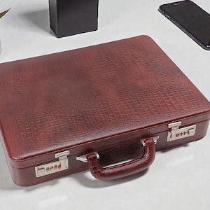 Puede incluir: Un maletín de cuero burdeos con un patrón texturizado similar al de un cocodrilo. El maletín tiene un cierre de combinación y un asa a juego. Una planta suculenta en una maceta blanca, una caja negra y un teléfono inteligente están en el fondo.