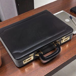 May include: A black leather briefcase with a combination lock and a black handle. The briefcase is on a wooden desk, next to a potted plant, a tablet, a pen, and a smartphone.