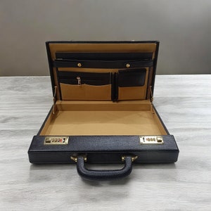 May include: Open black leather briefcase with a gold combination lock. The interior is tan with multiple pockets and compartments. The handle is black and the case is sitting on a light gray surface.