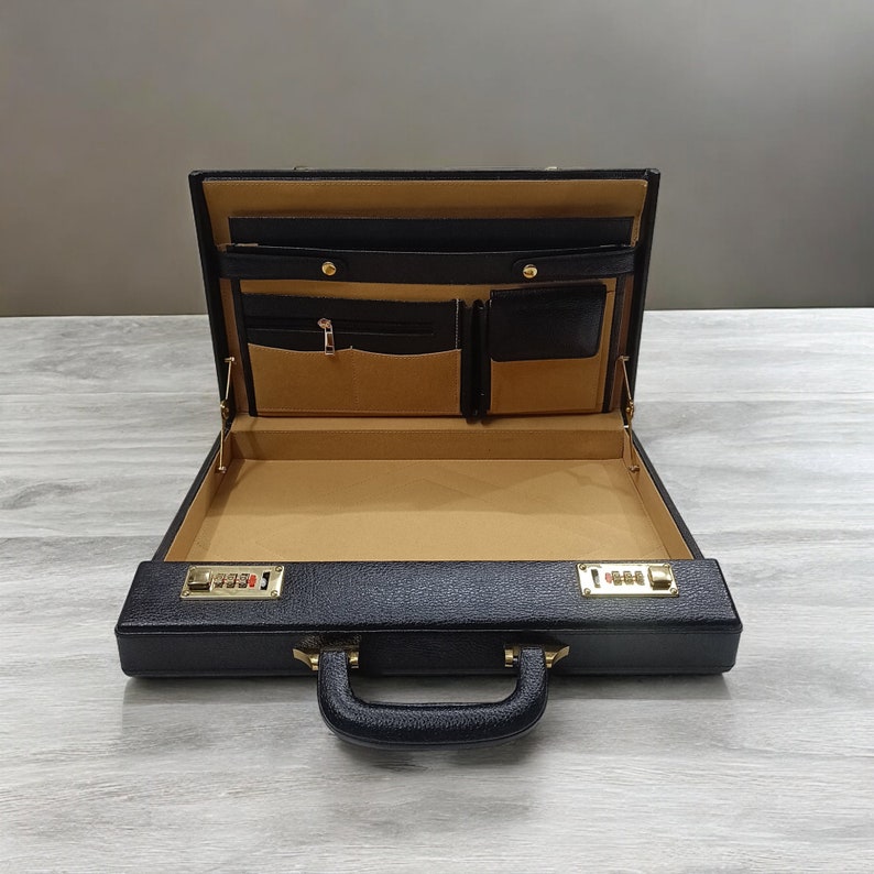 May include: Open black leather briefcase with a gold combination lock. The interior is tan with multiple pockets and compartments. The handle is black and the case is sitting on a light gray surface.
