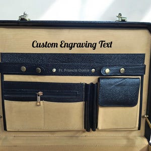 May include: Open black leather briefcase with multiple compartments and gold-colored hardware. The interior features a tan lining and the text "Custom Engraving Text". Includes a small text that reads "Fr. Francis Ouma".