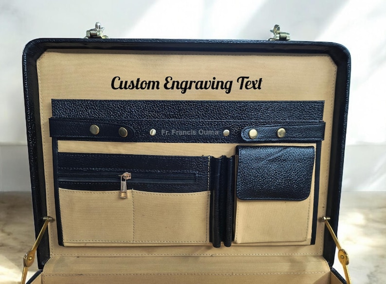 May include: Open black leather briefcase with multiple compartments and gold-colored hardware. The interior features a tan lining and the text "Custom Engraving Text". Includes a small text that reads "Fr. Francis Ouma".