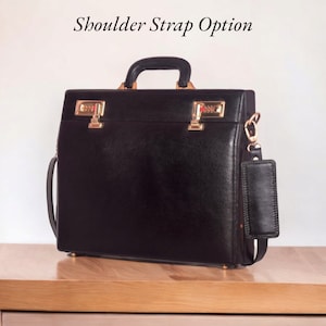May include: Black leather briefcase with gold-colored hardware, including two combination locks. Features a top handle and a detachable shoulder strap. The text "Shoulder Strap Option" is visible above the briefcase.