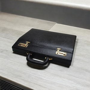 May include: A black leather briefcase with gold-colored combination locks and hardware. The briefcase has a handle and is closed. The surface has a textured appearance.