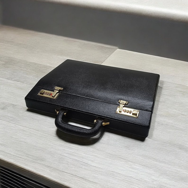 May include: A black leather briefcase with gold-colored combination locks and hardware. The briefcase has a handle and is closed. The surface has a textured appearance.