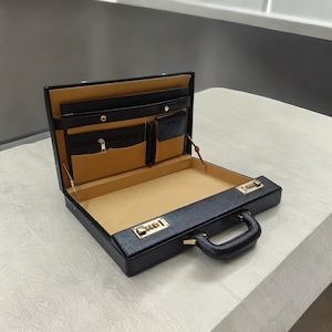 May include: Open black leather briefcase with a combination lock and a handle. The interior features tan lining, pockets, and compartments for organization. Gold-colored hardware accents the briefcase.